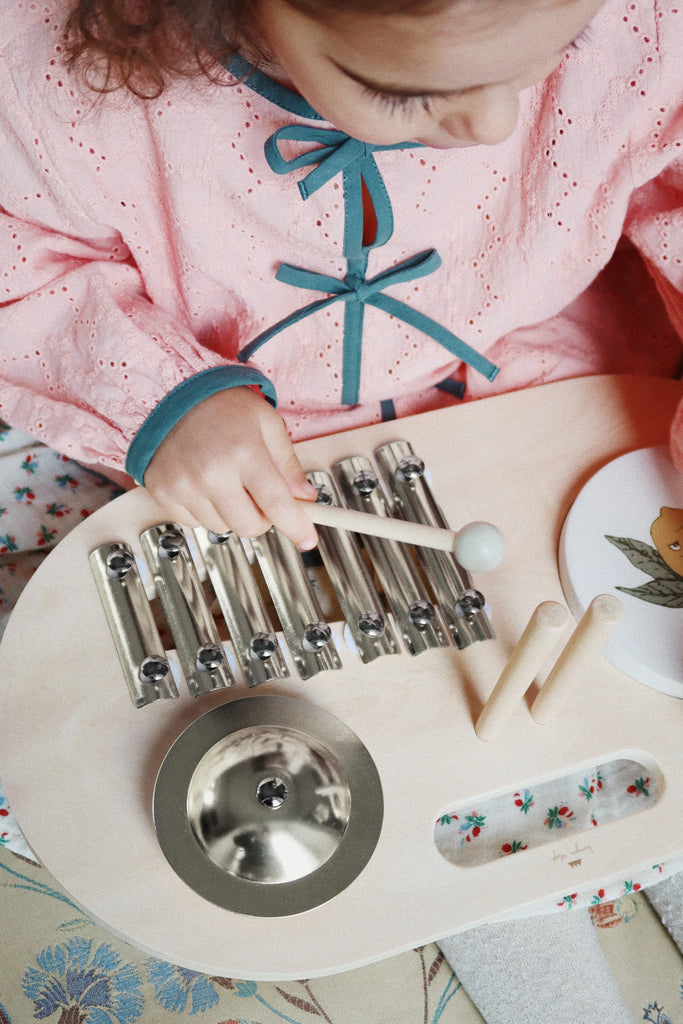 A child in a pink dress joyfully plays with the Konges Sløjd - Wooden Music Board, made from FSC-certified plywood, hitting its metal bars with a mallet. The toy includes a round metal cymbal and wooden sticks. The young musician is seated on a floral-patterned surface, captivated by the enchanting music.