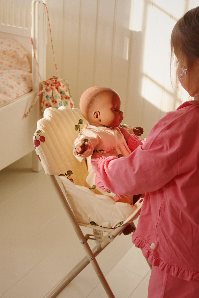 A young girl dressed in pink gently places a doll, complete with a doll accessory, in a Konges Sloejd High Chair - Cherry Glitter within a sunlit room. A crib and a floral bag are visible in the background.