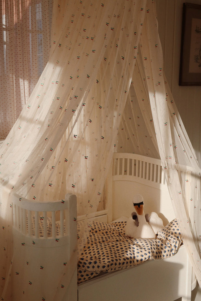 In a warmly lit room, a cozy baby crib creates a soft, serene atmosphere with the help of the Konges Sloejd Activity Music Swan gently playing Brahms' lullaby. The crib is adorned with delicate, patterned bedding and features a swan stuffed toy complementing its charm.