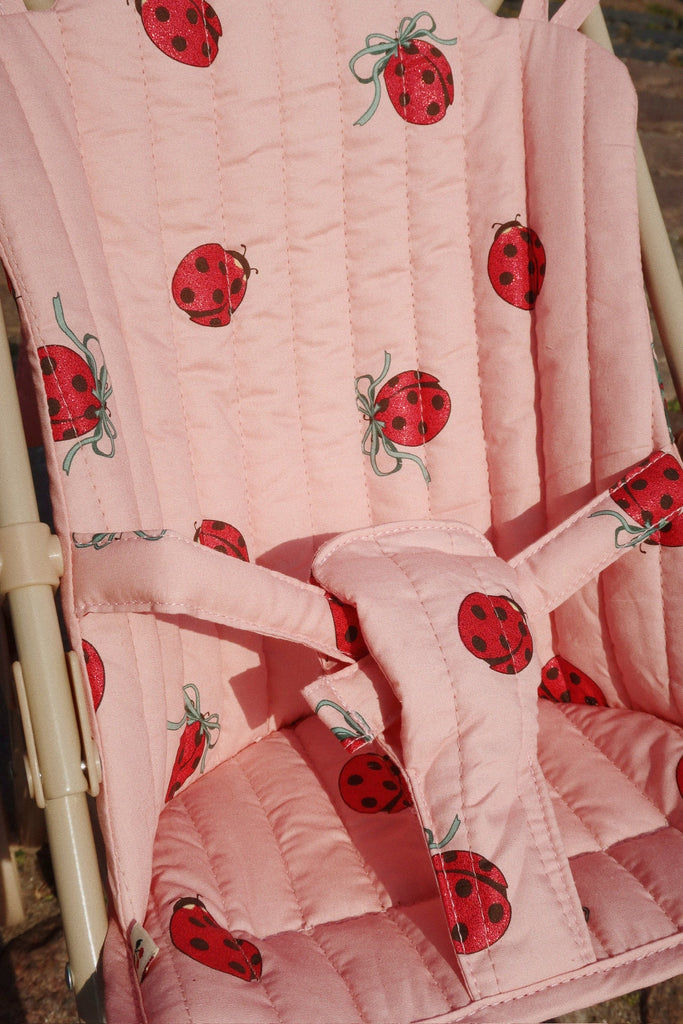 Close-up of the Konges Sloejd Doll Stroller seat cover, featuring pink quilted fabric with red ladybugs and green bows. The vertically stitched padding provides comfort and added stability for a child.