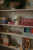 Wooden shelves with toys, colorful boxes, a ceramic cat, the Konges Sloejd Money Bank - Kitty, a row of colored markers, a toy train, miniature houses, and plants—all arranged neatly against a white paneled wall.