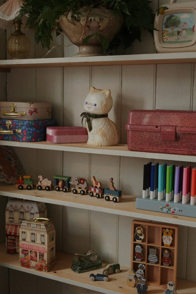 Konges Sloejd Wooden Animal Train, beechwood shelves with colorful boxes, a ceramic cat, markers, a model house, small cars, animal figurines, and a plant in a pot on the top shelf against a wooden wall.