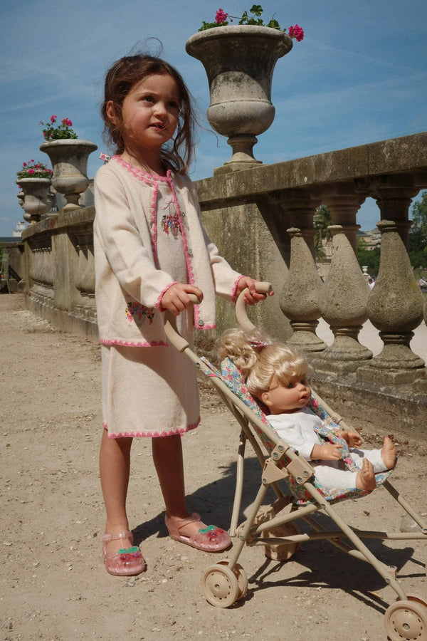 A young girl in a cream dress and pink sandals pushes the Konges Sloejd Doll Stroller - Fiola outdoors by a stone balustrade with potted pink flowers on a sunny day.