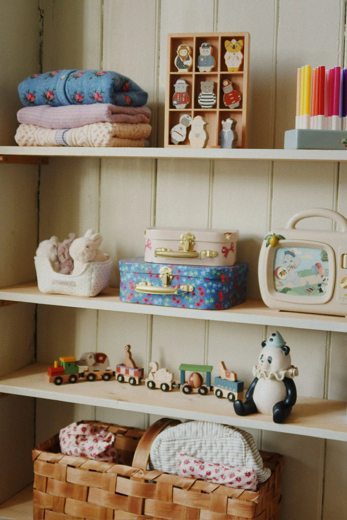 Konges Sloejd Wooden Animal Train with magnetic cars, folded blankets on wooden shelves, colorful suitcases, a toy radio, art supplies, wooden animal figurines, a toy bear, and wicker baskets—all neatly organized in a cozy, vintage style.