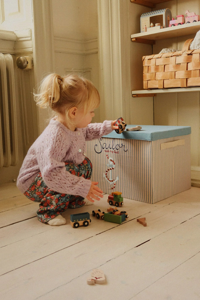 A young child with blonde hair, dressed in a lilac sweater and floral pants, crouches on a beechwood floor, playing with the Konges Sloejd Wooden Animal Train beside a striped storage box in a cozy, softly lit room.
