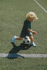 A young child with blond hair, dressed in a green outfit and white sneakers, is kicking a Konges Sloejd Football Set on a green turf field near a white line, enjoying some backyard practice.