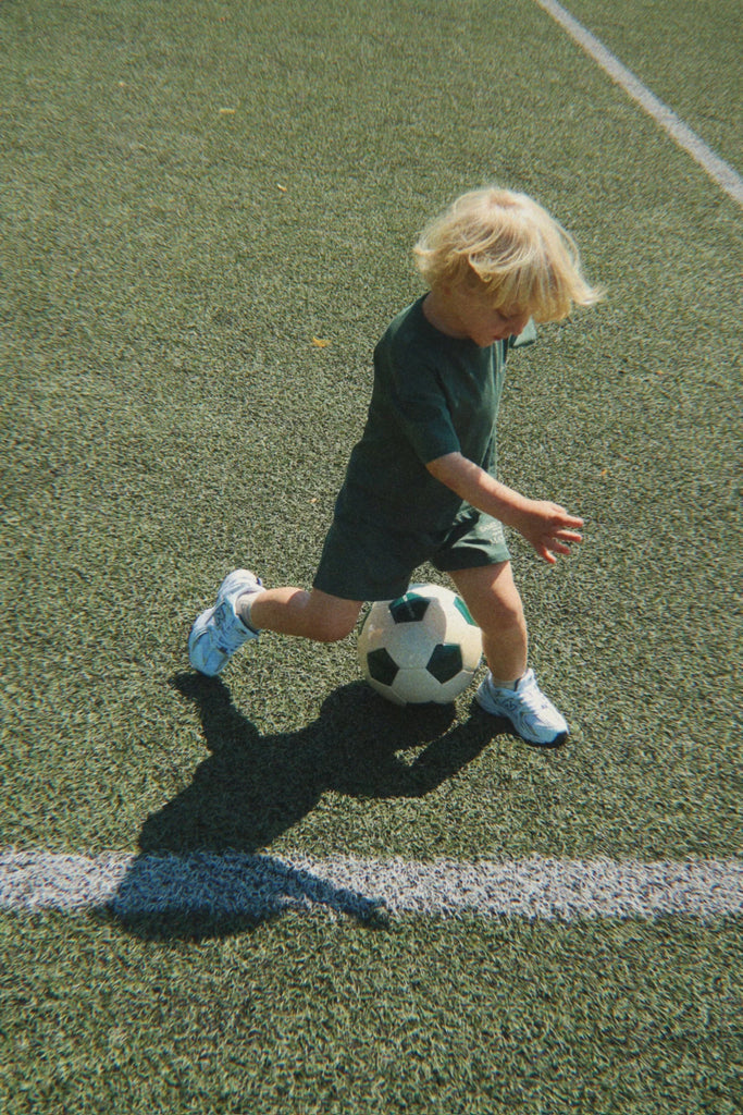 A young child with blond hair, dressed in a green outfit and white sneakers, is kicking a Konges Sloejd Football Set on a green turf field near a white line, enjoying some backyard practice.
