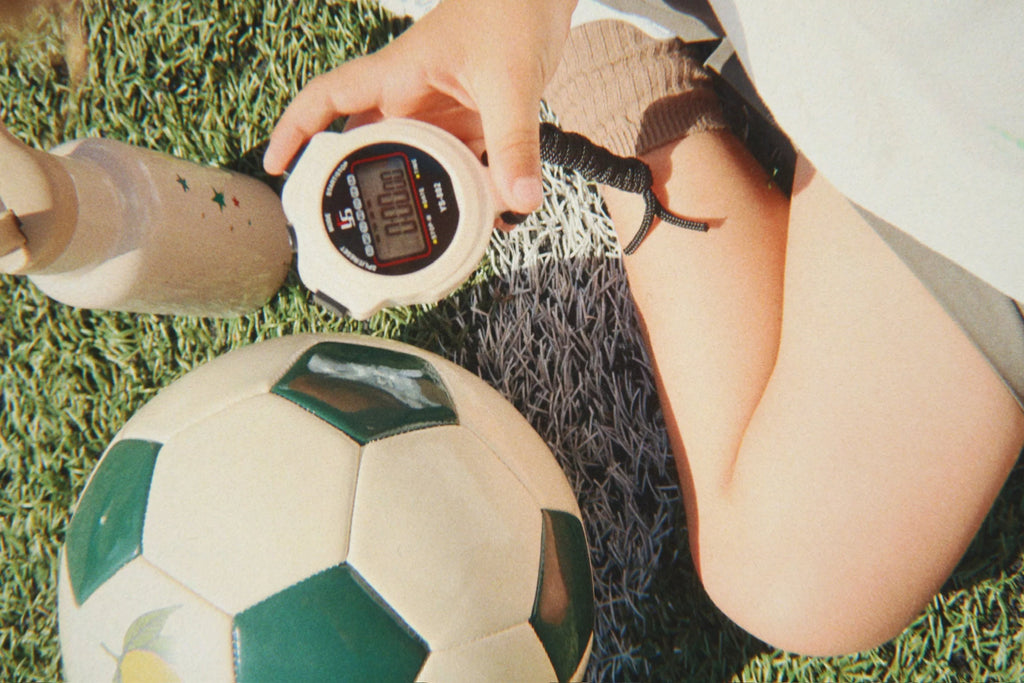 A child sits on the grass holding a stopwatch near their knees, beside a Konges Sloejd Football Set and a water bottle—suggesting a break during backyard practice or timing an activity with their football set.