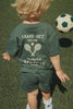 A young child with blonde hair, dressed in a green outfit that reads “Game Set You Are My Perfect Match,” walks on the grass near a Konges Sloejd Football Set, ready for some backyard practice.