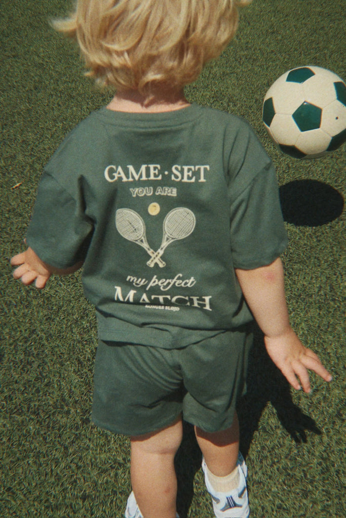 A young child with blonde hair, dressed in a green outfit that reads “Game Set You Are My Perfect Match,” walks on the grass near a Konges Sloejd Football Set, ready for some backyard practice.