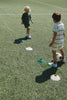 Two young children on a soccer field stand near marker cones. One child stands with the Konges Sloejd Football Set, while the other faces them. Both are dressed in casual clothes and sneakers, ready for some backyard practice.