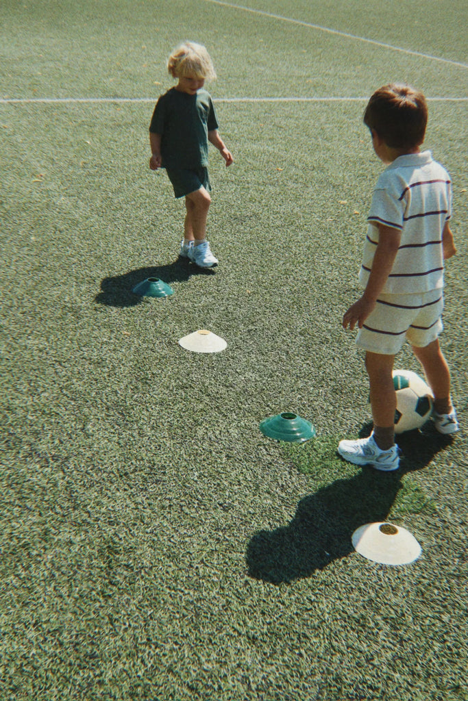 Two young children on a soccer field stand near marker cones. One child stands with the Konges Sloejd Football Set, while the other faces them. Both are dressed in casual clothes and sneakers, ready for some backyard practice.