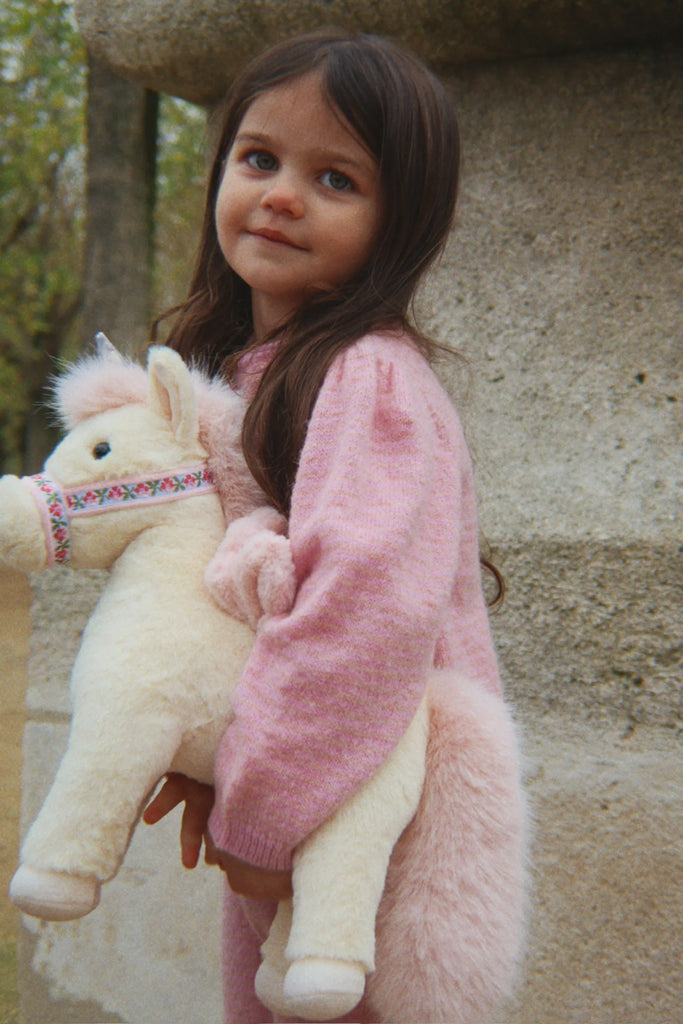 A young girl with long brown hair, dressed in a pink sweater, stands outdoors holding the Konges Sloejd Starlight Unicorn Stuffed Animal, featuring pink wings and a colorful harness. She gazes slightly upward with a calm expression, while stone structures and trees appear in the background.