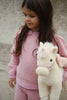A young child in a pink sweater holds the Konges Sloejd Starlight Unicorn Stuffed Animal, featuring soft pink wings. The child, with long brown hair, stands against a neutral textured background and gazes slightly to the side with a calm expression.