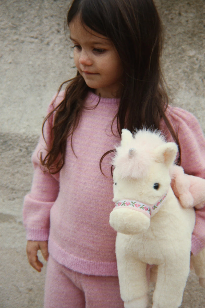 A young child in a pink sweater holds the Konges Sloejd Starlight Unicorn Stuffed Animal, featuring soft pink wings. The child, with long brown hair, stands against a neutral textured background and gazes slightly to the side with a calm expression.
