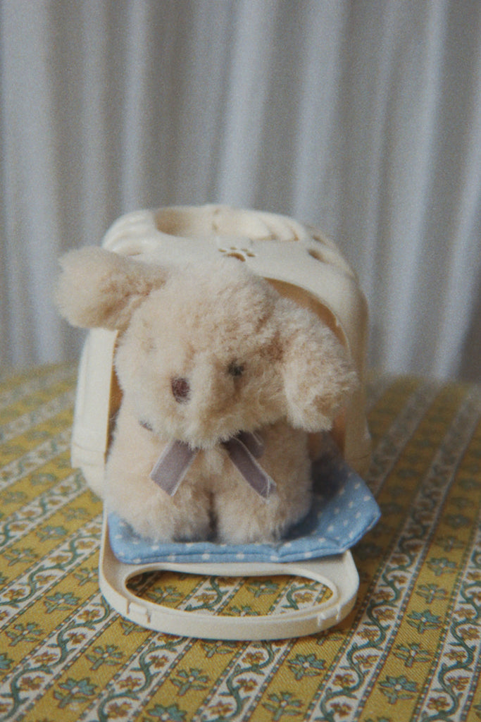 A Konges Sloejd Take Care of Me Animals - Dog plush, small and fluffy with floppy ears, sits on a blue polka-dot blanket in front of a white plastic carrier on a yellow patterned surface, with a white curtain in the background—waiting for you to take care of it.