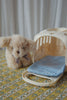 A plush dog from the Konges Sloejd Take Care of Me Animals collection—adorned with a purple ribbon—sits beside a cream-colored pet carrier featuring a blue polka-dot cushion, displayed on a patterned surface in front of white curtains, inviting you to care for it.