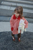 A young child wearing a pink coat and brown shoes stands on a city crosswalk, holding the Konges Sloejd Starlight Unicorn Stuffed Animal.