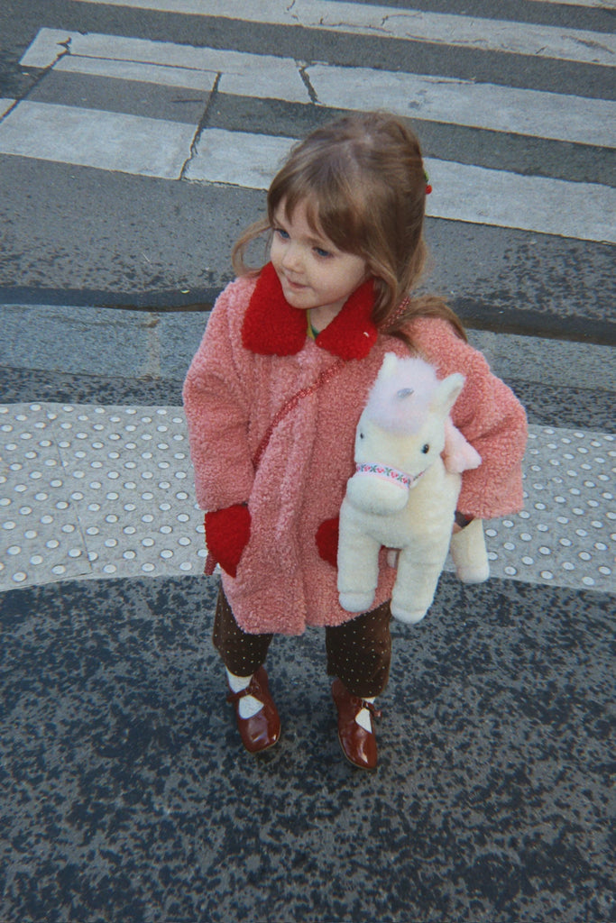 A young child wearing a pink coat and brown shoes stands on a city crosswalk, holding the Konges Sloejd Starlight Unicorn Stuffed Animal.