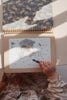 A child is coloring a picture of a unicorn riding a broomstick in the "My Magic Water Book - Off White." The child's hand is holding a black marker, and the spiral-bound book features reusable pages with blue and white patterns. The child, dressed in a ruffled, long-sleeved dress, enjoys every moment.