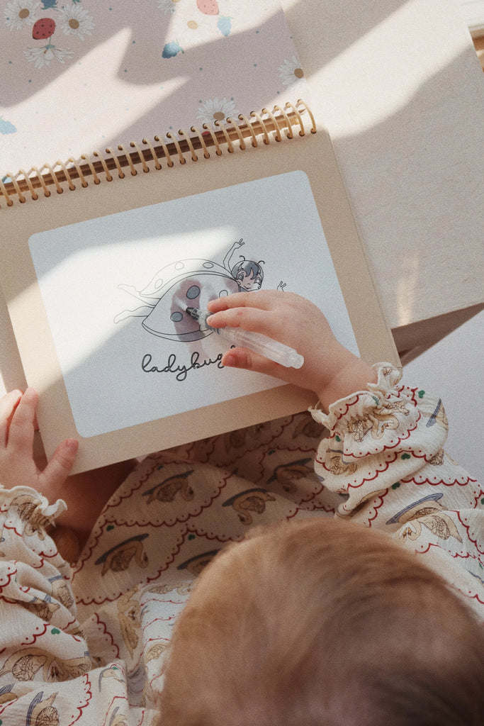 A young child, dressed in a patterned outfit, is drawing on a "My Magic Water Book - Off White" with reusable pages. The drawing depicts a ladybug, labeled "ladybird." The child is holding a white crayon or marker and is focused on the water book. Background elements include floral designs.