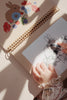 A child with a floral-patterned sleeve uses My Magic Water Book - Off White to color in a bunny illustration on a spiral-bound sketchbook. Another reusable page with a colorful drawing of a bunny and flowers lies beneath. Soft natural light adds a warm tone to this perfect travel activity scene.