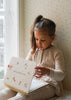 A young girl with a ponytail is sitting by a window, wearing a beige sweater dress. She is looking down at an open My Magic Water Book - Sleet. The room has light-colored wallpaper with small patterns, and natural light is streaming through the window.