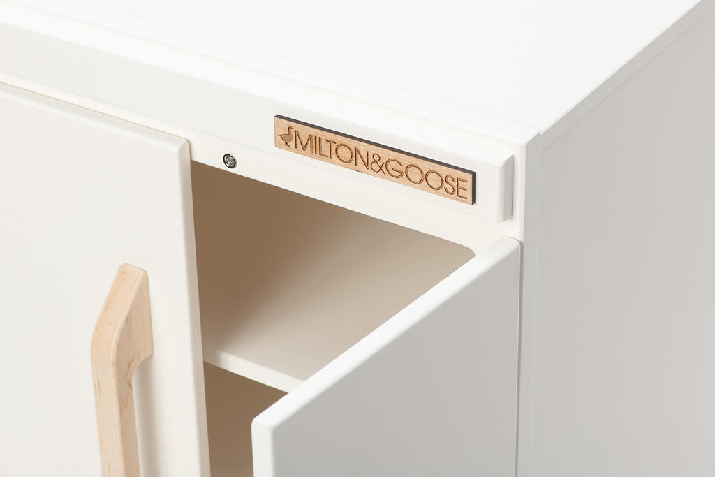 A close-up of the Milton & Goose Essential Play Refrigerator—featuring a white finish, wooden handle, open door, and top label—made in the USA and ideal for imaginative play or storing pretend food.