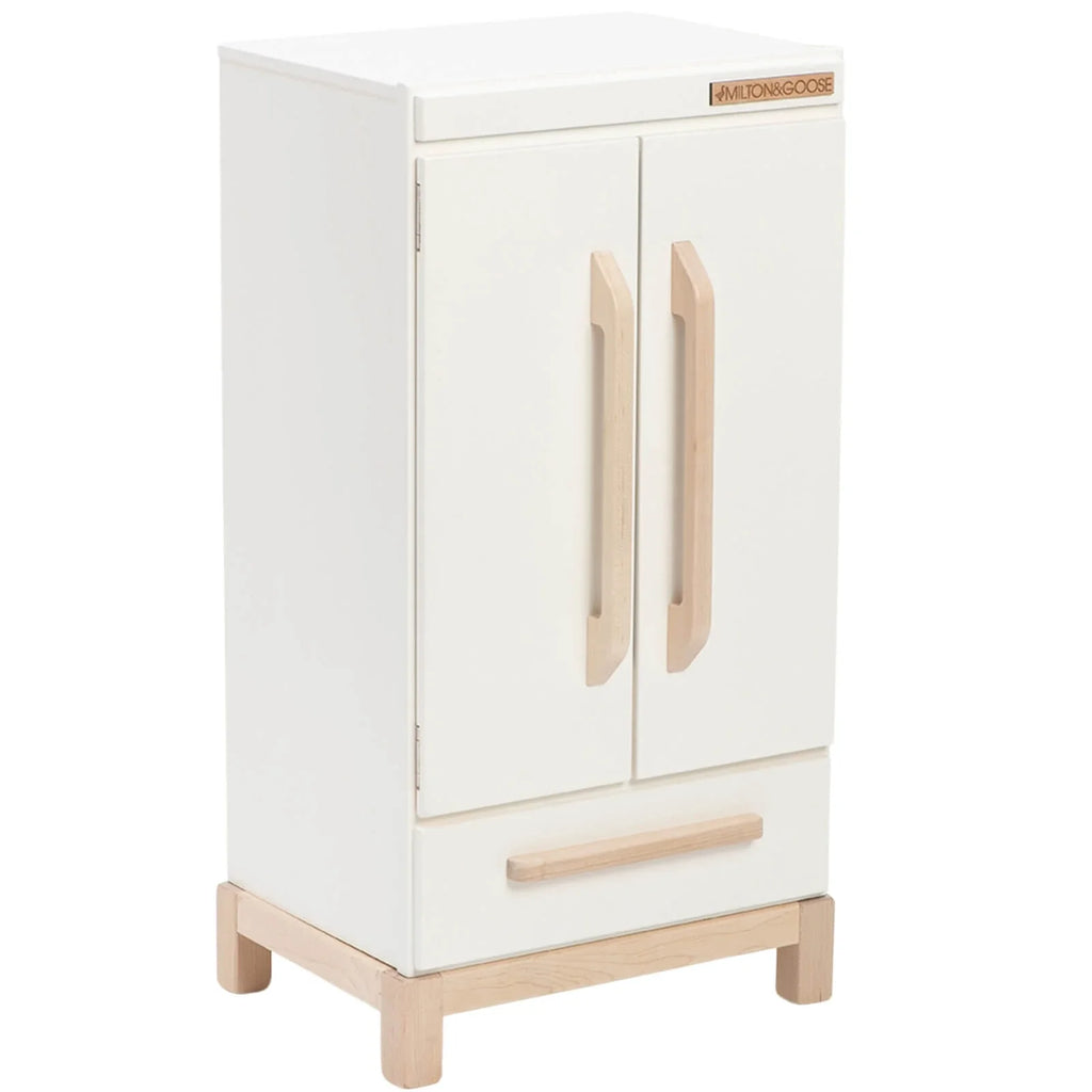 The Milton & Goose Essential Play Refrigerator is a white wooden cabinet with two doors, light wood handles and legs, a lower drawer, minimalist design, and a small brand label—perfect for play kitchens or imaginative play. Made in the USA.