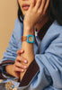 A woman in a light blue jacket rests her chin on her hand, displaying the Mini Kyomo Watch - Havana Sky with a tan strap on her wrist. Her face is partially out of the frame.