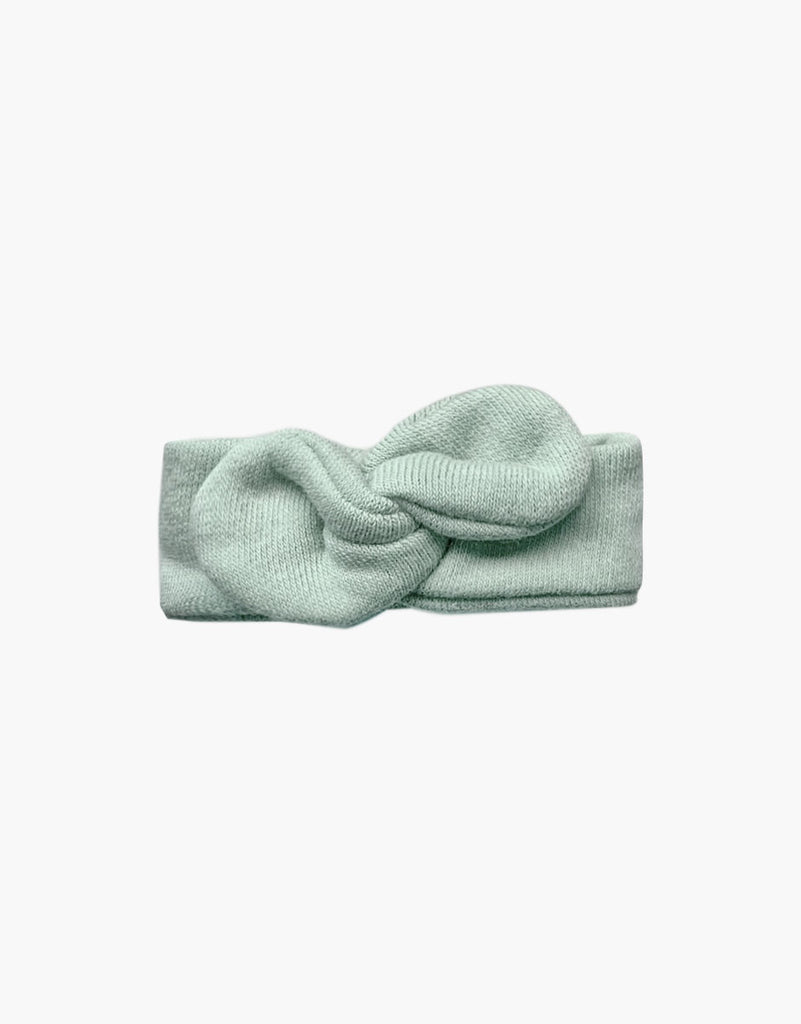 A soft, pale green, knitted headband with a twisted knot design in the center, perfect for accessorizing Minikane dolls, shown against a plain white background: Minikane Doll Clothing | Green Tea Doll Headband.
