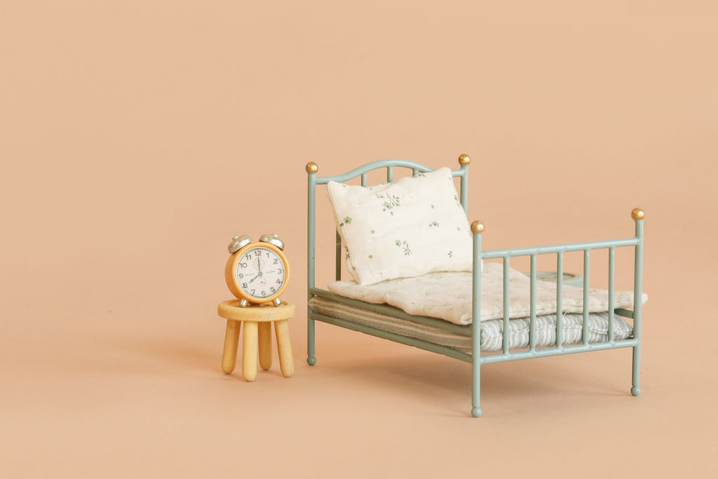 A small metal bed with pillows and a blanket sits next to the Maileg Stool Set, Mouse in Light Rose/Light Mint, which holds a silver alarm clock—all set against a beige background.