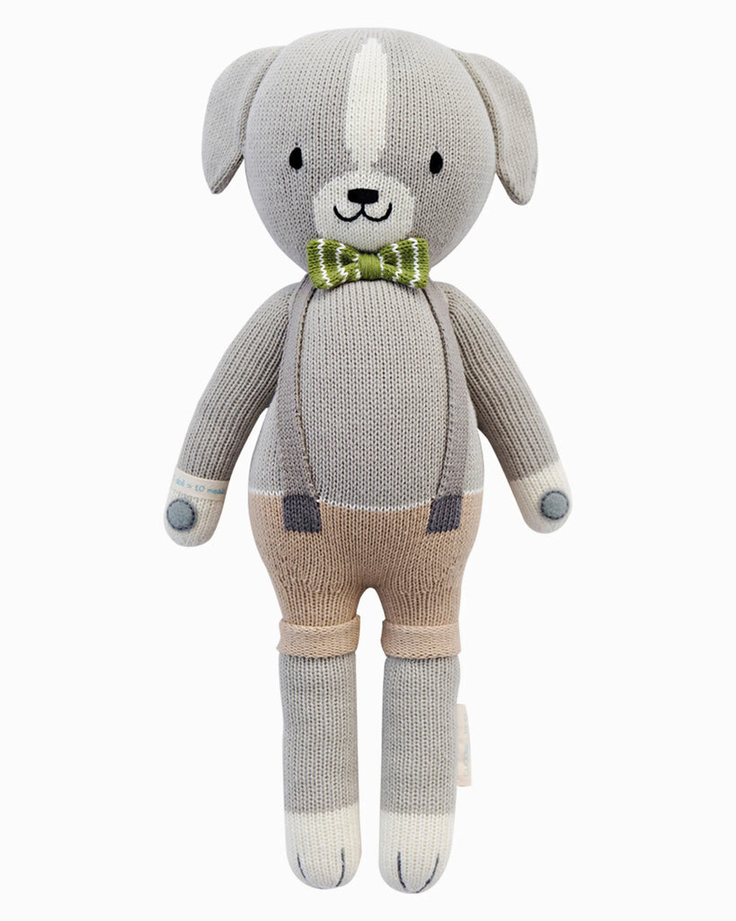Cuddle + Kind Noah the Dog is a fair trade, hand-knit gray dog doll with floppy ears, beige shorts, suspenders, a green plaid bow tie, and white accents on its paws and forehead. It features a friendly, simple face.