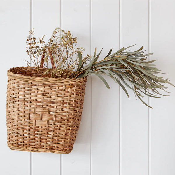 The Rattan Bowery Basket hangs elegantly on a white paneled wall, blending decor and storage perfectly, with dried flowers and greenery gracefully spilling over the top.