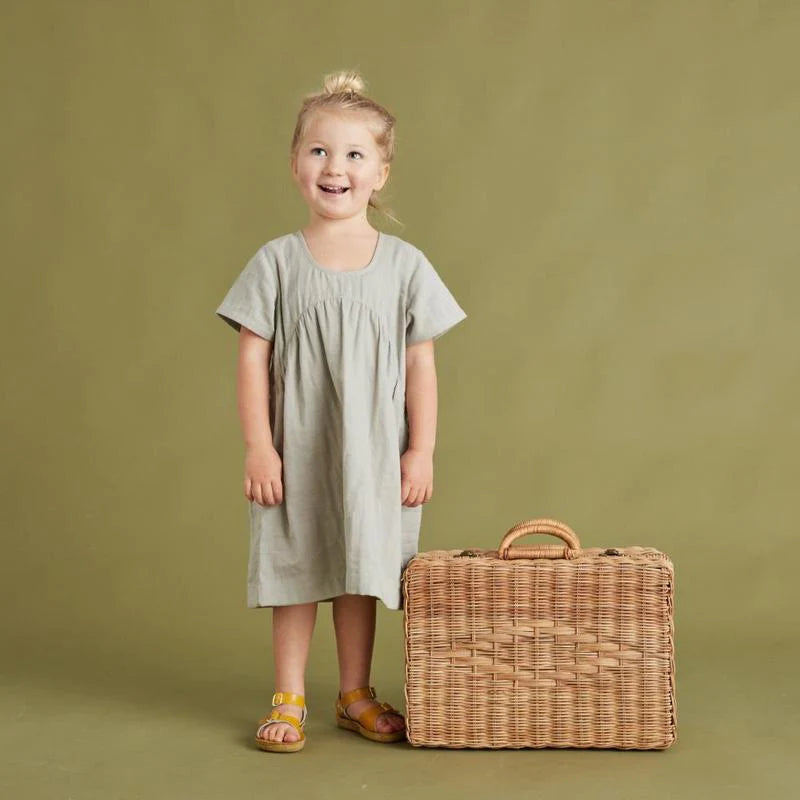 A young girl with light blonde hair stands smiling beside a large Easter Basket Set trunk, wearing a gray dress and colorful sandals against a green backdrop.