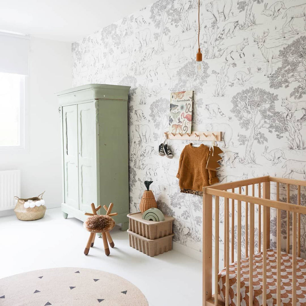 A serene nursery featuring a sage green wardrobe, a whimsical forest-themed wallpaper, a wooden crib, Faux Bambi chairs, and various other items like toys and a hanging sweater.