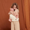 A young child stands against a brown backdrop, smiling gently while hugging a pink stuffed animal carried in the Olli Ella Doll Carrier with a rainbow print.