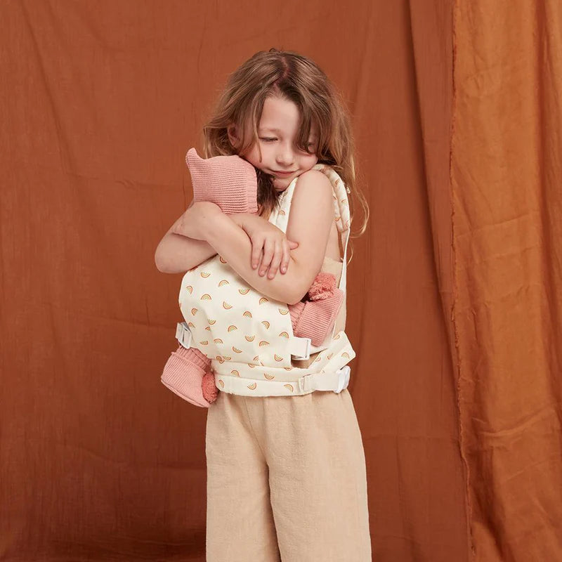A young child stands against a brown backdrop, smiling gently while hugging a pink stuffed animal carried in the Olli Ella Doll Carrier with a rainbow print.