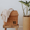 The Olli Ella Hanging Rattan Basket Hutch, resembling a small house with a window, sits on a wooden stool. A light-colored knit blanket is draped over the basket. A tall potted plant with large green leaves stands next to the stool against a white background. This charming handcrafted rattan storage solution blends functionality and style effortlessly.