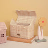 An Olli Ella Casa Bag, featuring a hand-woven rattan design and open lid, rests on a closed book beside a decorative wooden flower with a yellow blossom and red center, all set against a beige background.