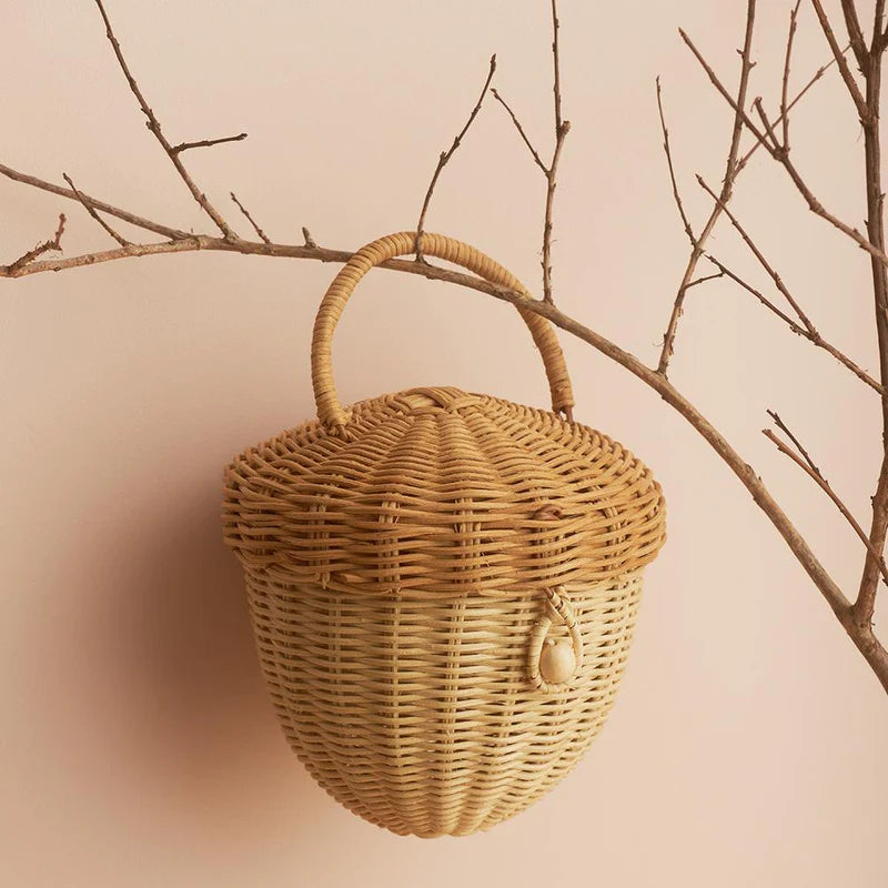 Olli Ella Rattan Acorn Bag with a lid and handle, hanging on a hook beside bare tree branches against a soft peach-colored wall.