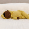 An Olli Ella Dozy Dinkum - Mini soft cotton doll with brown skin, closed eyes, and black hair lies on a white quilt. The baby toy wears a yellow hooded onesie with buttons and a small embroidered rainbow on the chest.