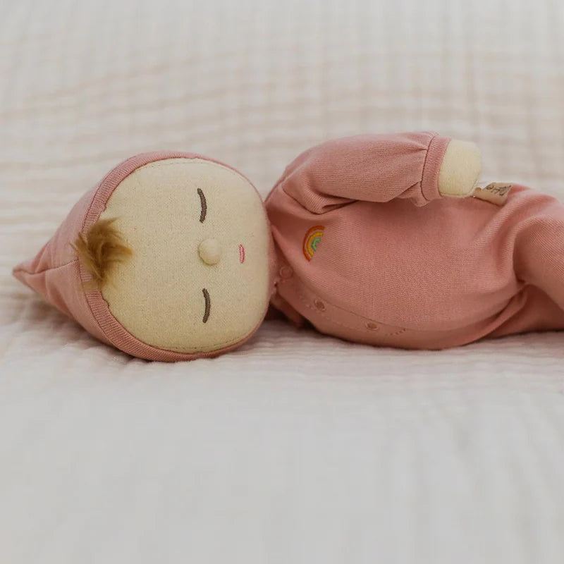 Olli Ella Dozy Dinkum - Moppet is a soft cotton baby doll with a pink outfit and hat, closed eyes, a brown hair tuft, and a stitched rainbow on its chest, shown resting on a light textured blanket.