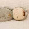 The Olli Ella Dozy Dinkum - Pickle is a soft cotton doll with closed embroidered eyes, light brown hair, and a beige face, dressed in a light gray outfit with a rainbow patch—perfect as a baby's first toy.