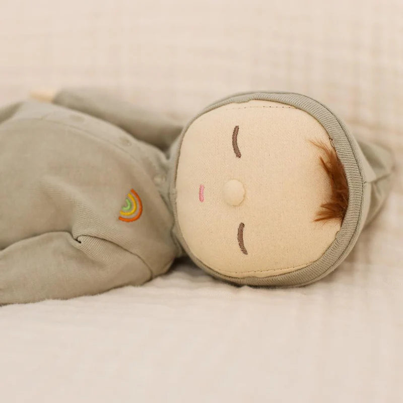 The Olli Ella Dozy Dinkum - Pickle is a soft cotton doll with closed embroidered eyes, light brown hair, and a beige face, dressed in a light gray outfit with a rainbow patch—perfect as a baby's first toy.