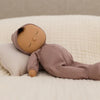 The Olli Ella Dozy Dinkum - Pip is a soft cotton doll with brown skin, closed eyes, and a mauve outfit and cap, resting on a cream pillow—perfect as a baby’s first toy.