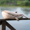 The Bukowski - Olympus Whale, a cuddly plush toy with a fuzzy underside, rests on a wooden railing near a calm lake, with greenery visible in the background.