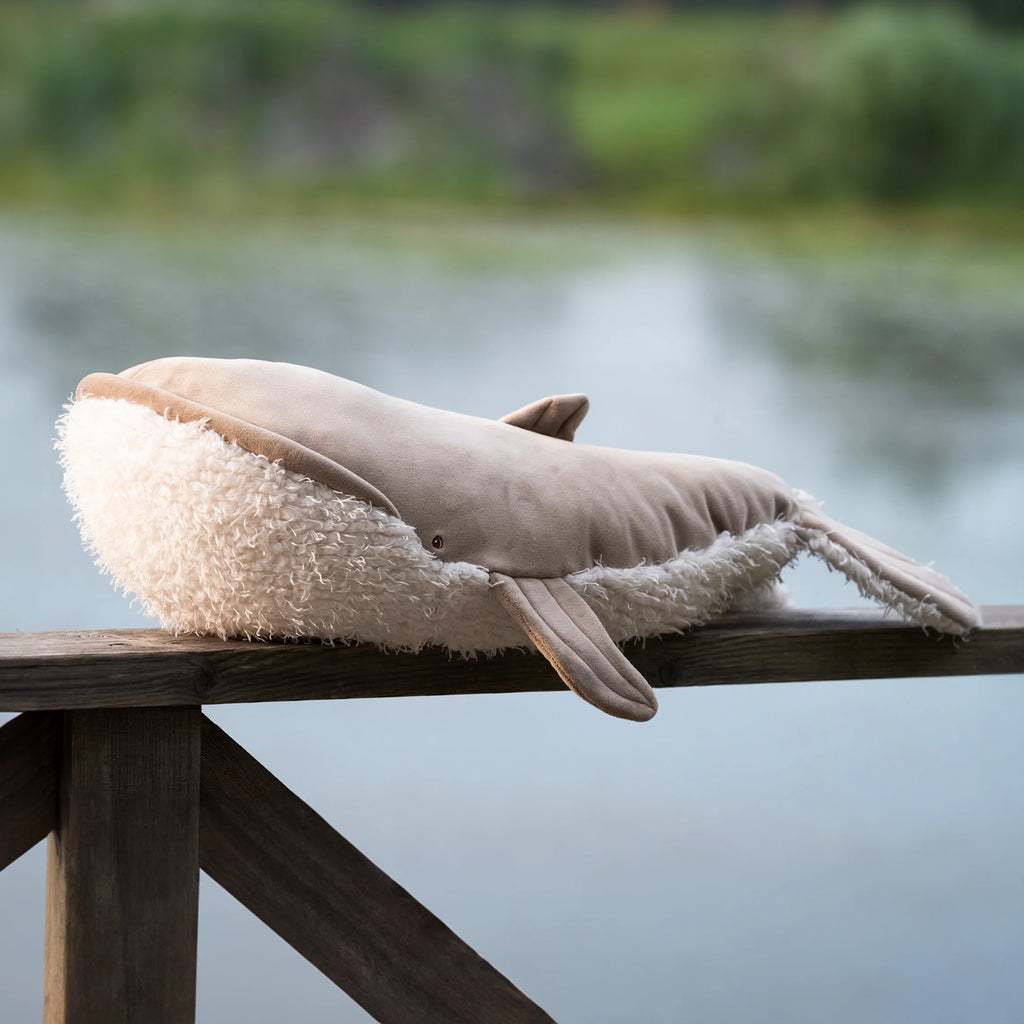 The Bukowski - Olympus Whale, a cuddly plush toy with a fuzzy underside, rests on a wooden railing near a calm lake, with greenery visible in the background.