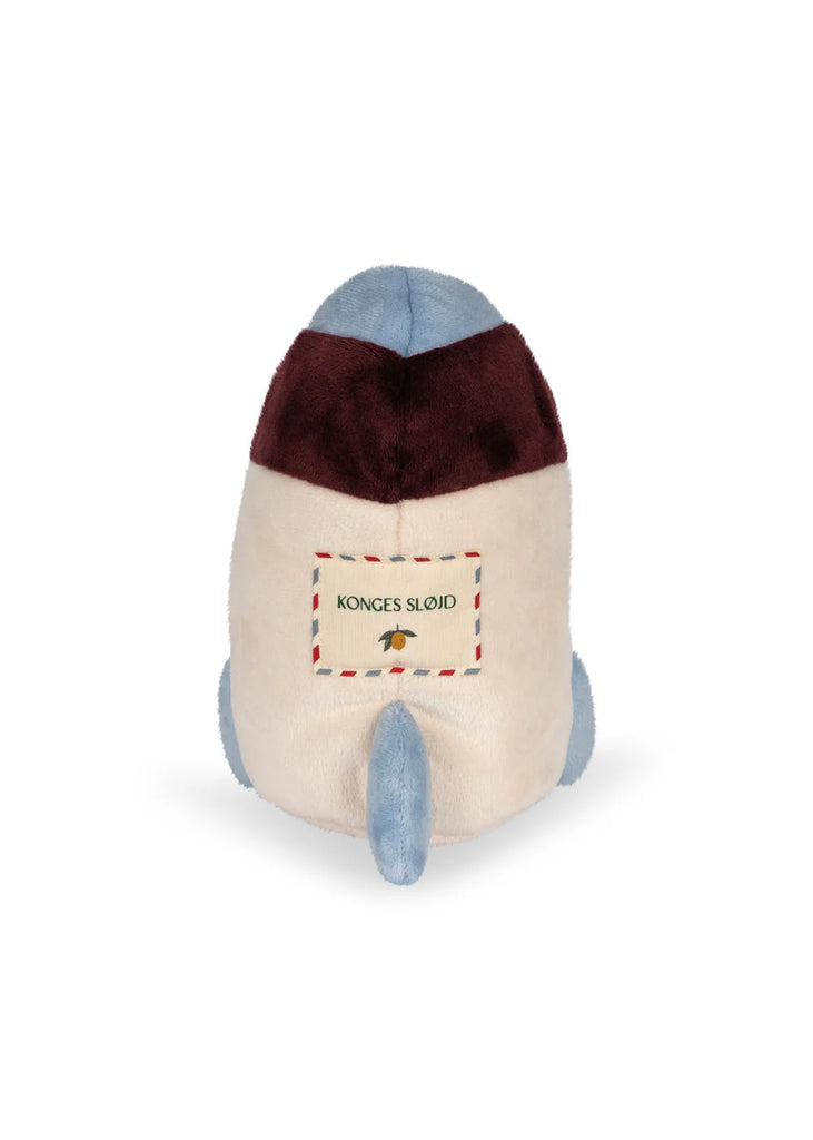 The Teddy In Outer Space Stuffed Animal - Rocket is a plush toy designed to resemble a cartoonish rocket, primarily white with blue and burgundy accents. It features a rectangular cream-colored label on one side that reads "Konges Slojd" and includes a small illustration of a bee. The toy is filled with recycled polyester.