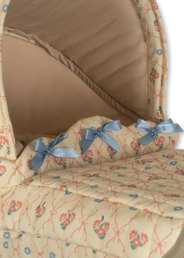 The Doll Pram - Fleur Decor, which ships in approximately two weeks, showcases a vintage-style charm with its soft, floral-patterned Moses basket in beige and pink. It features a cushioned interior adorned with three small blue bows on the side and is complemented by an organic cotton mattress.